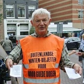 Toon borst, de muntenman, met twee papiertjes in zijn handen en een oranje hesje aan met daarop tekst om geld in te zamelen.