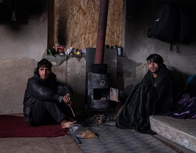 Portretfoto van twee vluchtelingen die op de grond zitten bij een ketel om zichzelf warm te houden in Bosnië