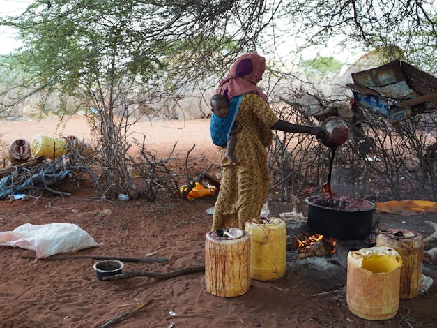 Vrouw giet rode vloeistof in pan met baby in draagzak op haar rug in Kenia