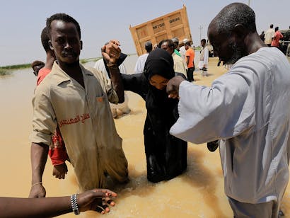 Twee mannen helpen een vrouw door het overstroomde gebied heen in Soedan