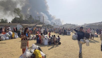 Grote groep vluchtelingen met spullen staat bij elkaar met grote rookwolken op de achtergrond