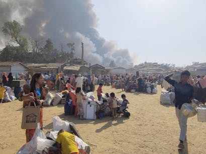 Grote groep vluchtelingen met spullen staat bij elkaar met grote rookwolken op de achtergrond