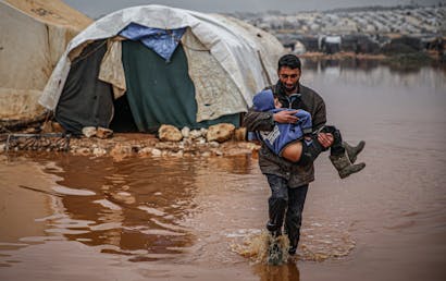 een man uit syrie draagt een kind door een overstroomd vluchtelingenkamp in idlib