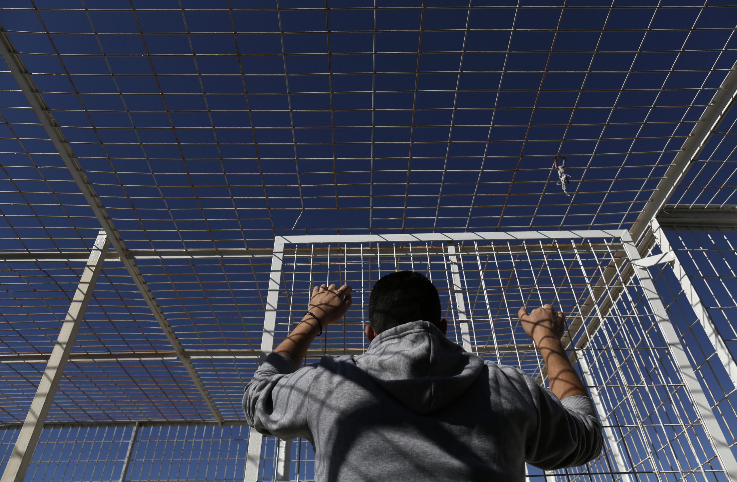 Een Syrische vluchteling staat met zijn rug naar de camera en houdt de tralies vast van een detentiecentrum in Griekenland, waar hij wordt vastgehouden.