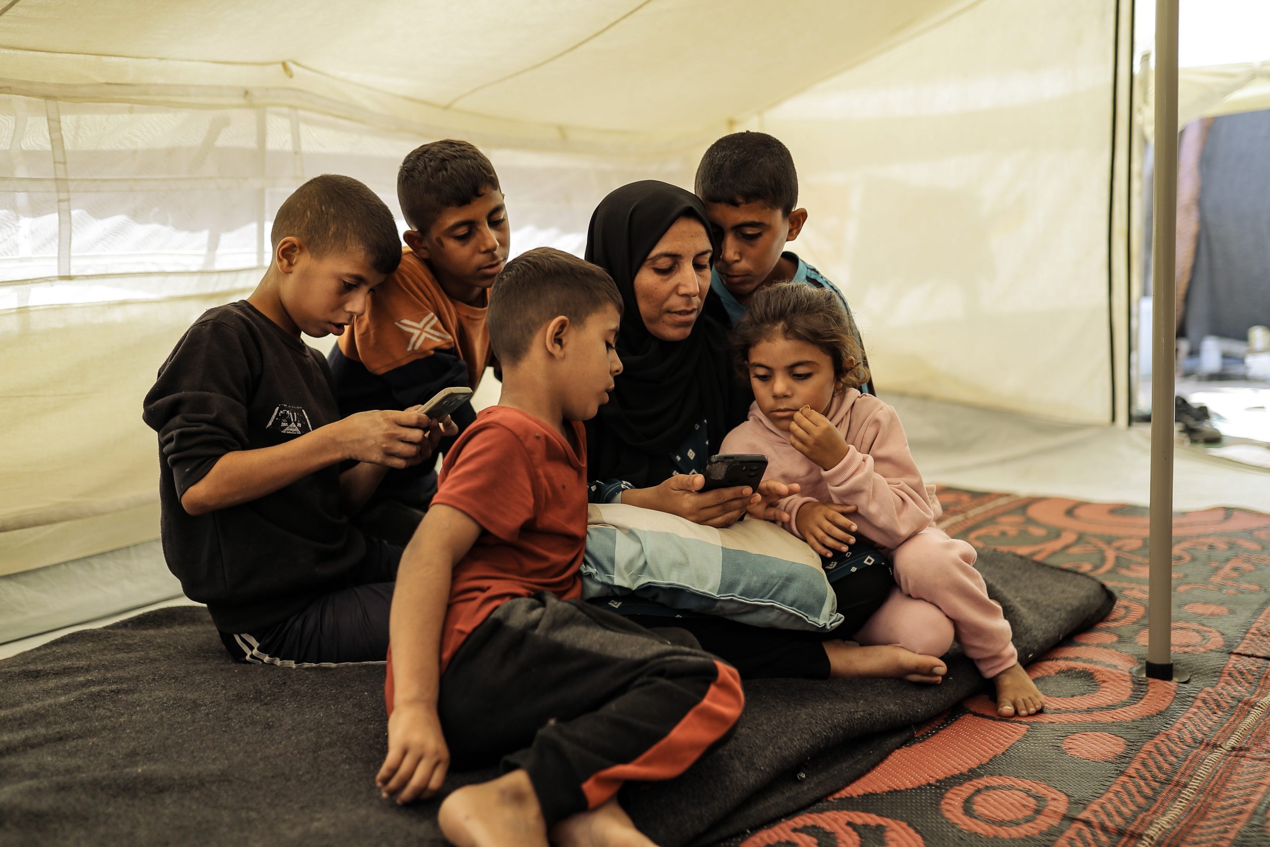 Amani en haar vijf kinderen zitten in hun tent in Gaza en kijken samen naar iets op een mobiele telefoon.