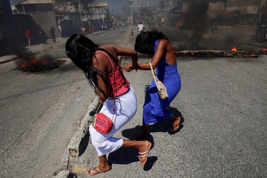 twee vrouwen rennen door een straat in Haiti, op de achtergrond is rook te zien.