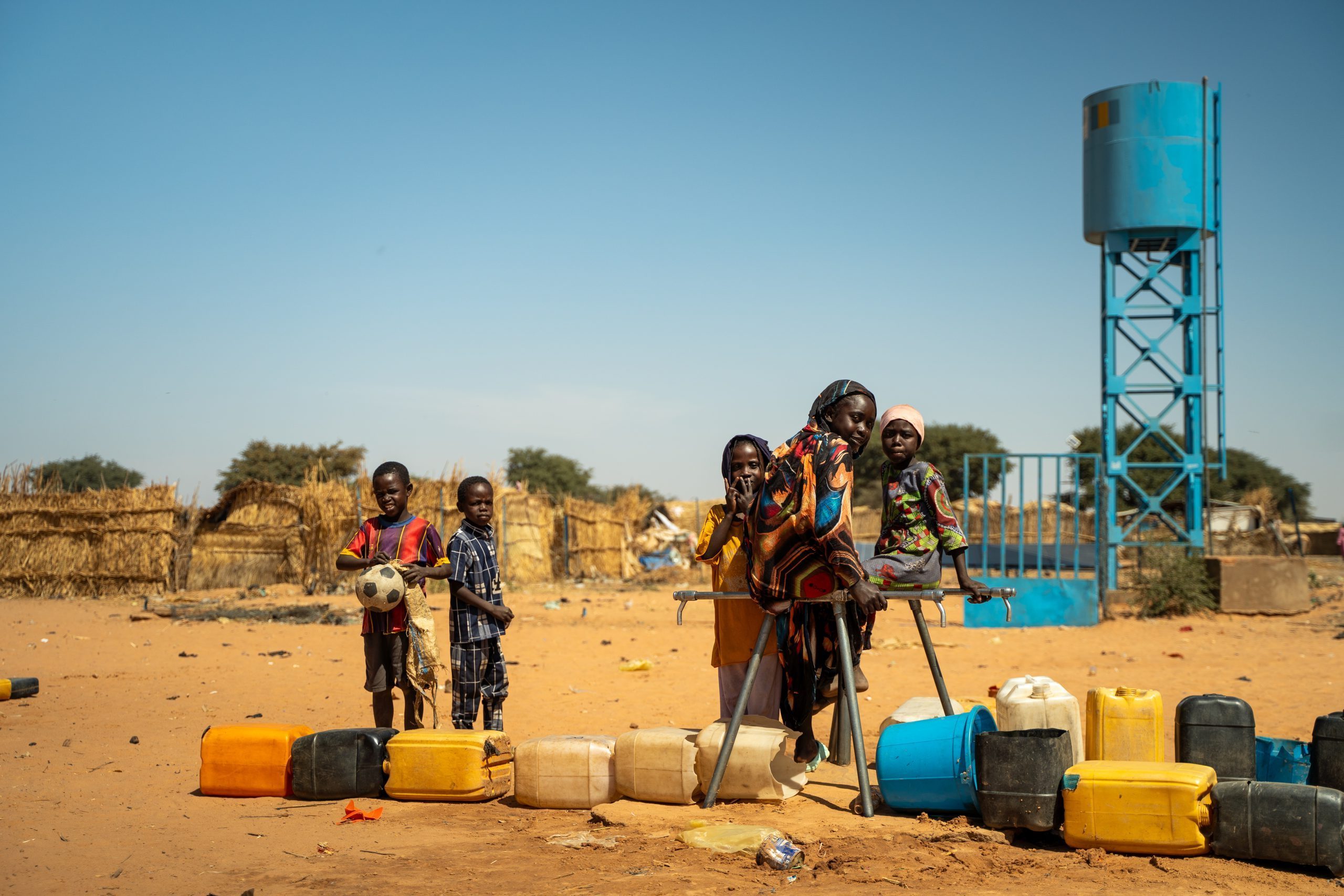 een groep kinderen staat voor het Adré vluchtelingenkamp in Tsjaad. Voor hen liggen jerrycans.