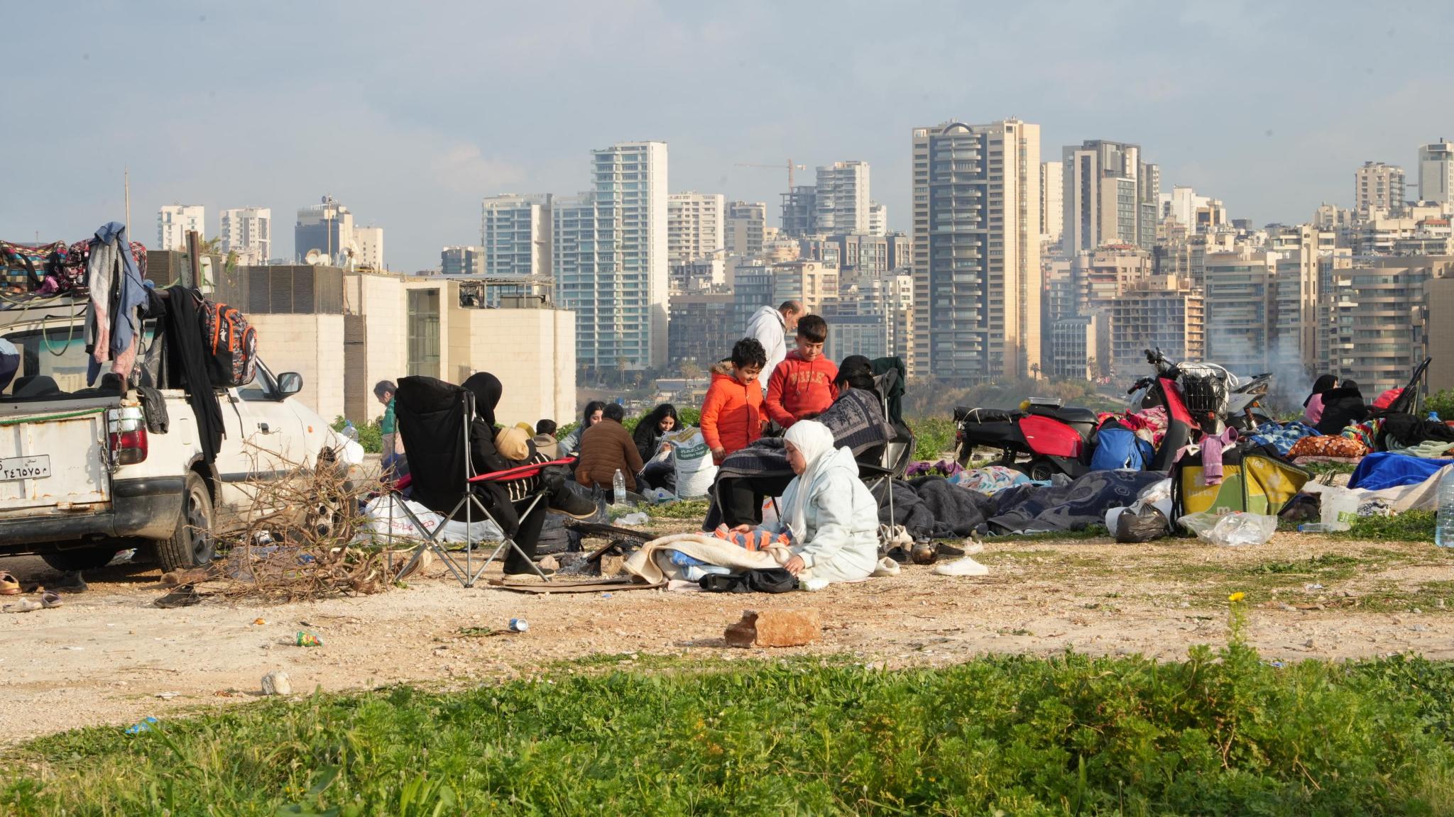 Een groep gevluchte Libanezen zit op de grond naast een auto, achter hen zie je Beiroet